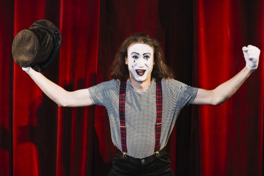 happy-mime-standing-front-red-curtain-holding-hat-flexing-his-muscle